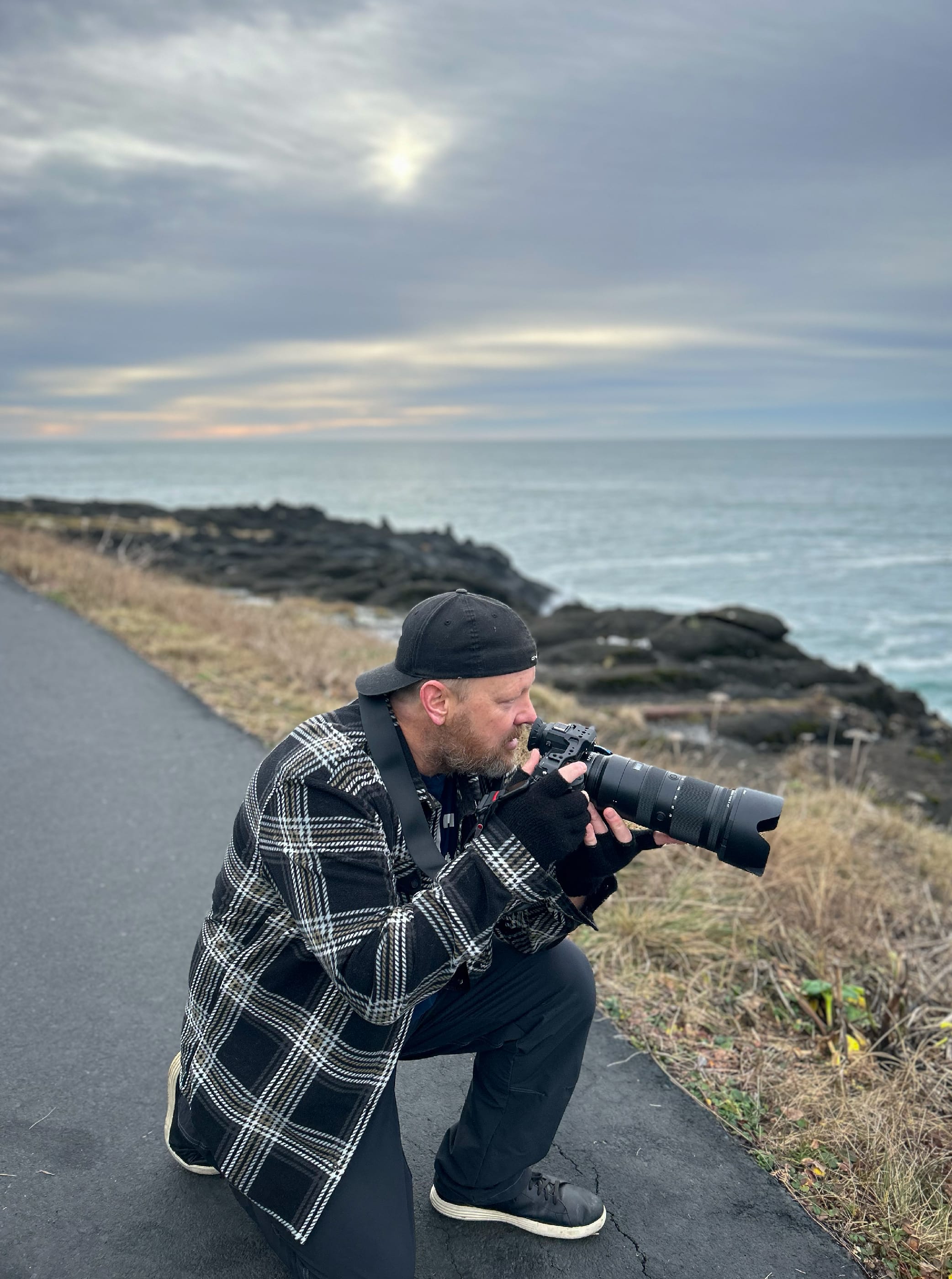 Jeremy Burke crouched at the edge of a paved coastal overlook with a long telephoto lens, photographing the surf below at dusk on the Oregon Coast.