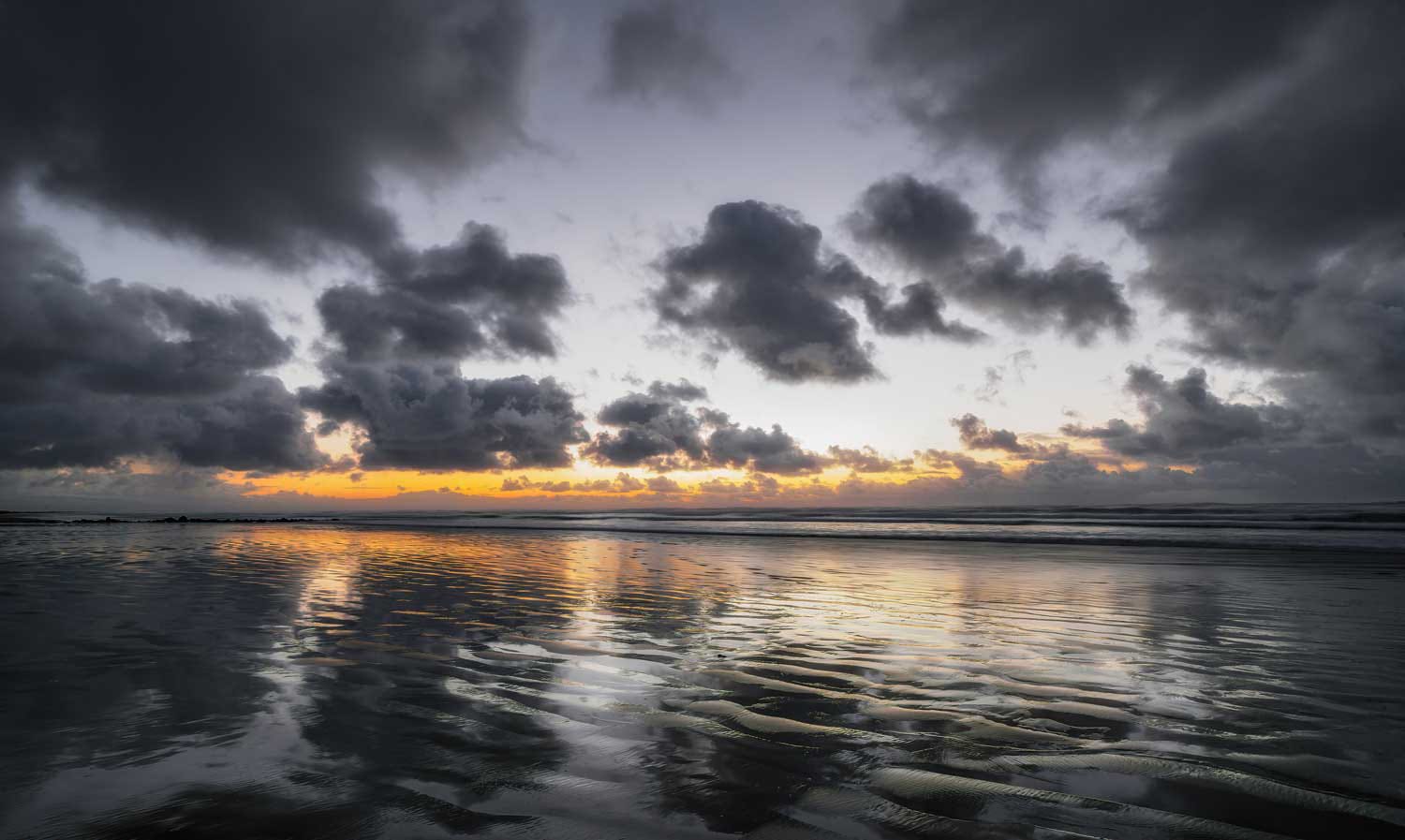 Low sun breaking beneath a heavy cloud deck and reflecting in ribbed wet sand at low tide, Newport, Oregon, at sunset.
