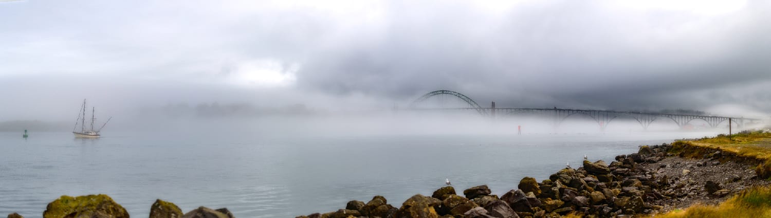 A single masted sailboat lying at anchor in fog on Yaquina Bay, Newport, Oregon, with the bay bridge dissolving into the weather behind it and wet riprap in the foreground.
