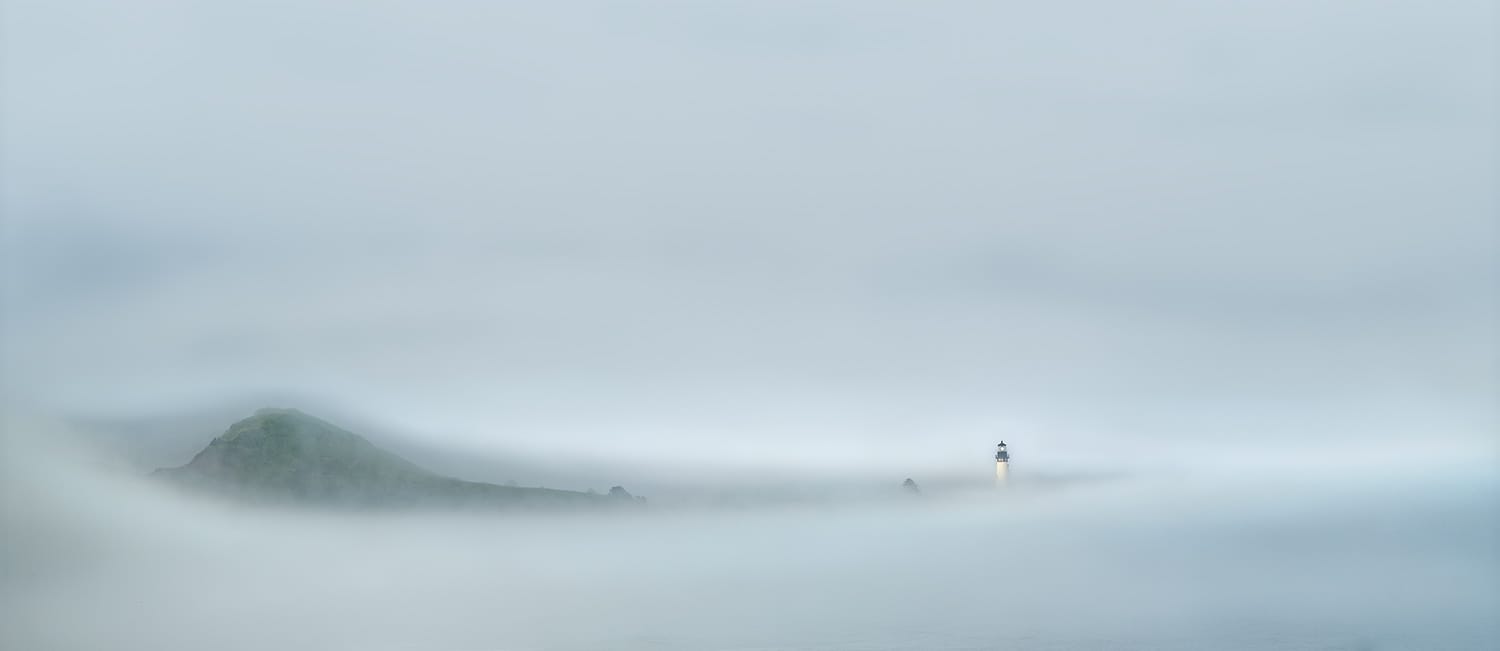 A lighthouse tower and the headland beneath it reduced to faint silhouettes inside a low bank of coastal fog, above a flat and windless sea on the Oregon Coast at first light.