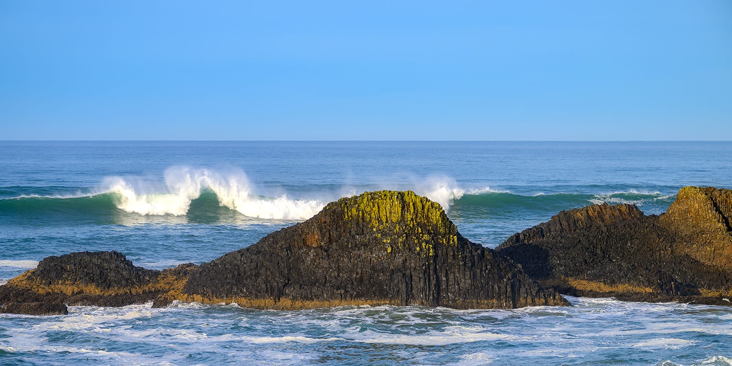 A single wave breaking white behind lichen-covered basalt reefs at Seal Rock, Oregon, under hard low sunlight and a clear blue sky.